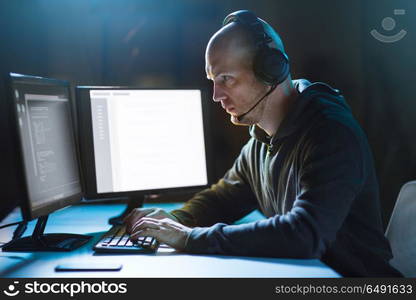 cybercrime, hacking and technology concept - male hacker with headset and coding on computer screen wiretapping or using computer virus program for cyber attack in dark room. hacker with computer and headset in dark room. hacker with computer and headset in dark room