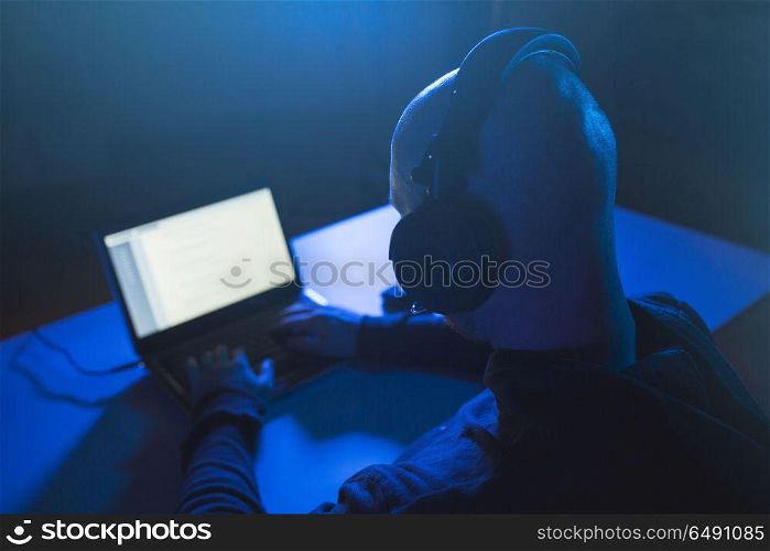 cybercrime, hacking and technology concept - male hacker with headset using laptop computer for cyber attack or wiretapping in dark room. hacker with headset typing on laptop in dark room. hacker with headset typing on laptop in dark room