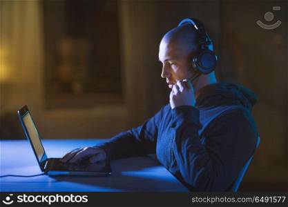 cybercrime, hacking and technology concept - male hacker with headset using laptop computer for cyber attack or wiretapping in dark room. hacker in headset typing on laptop in dark room. hacker in headset typing on laptop in dark room