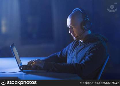 cybercrime, hacking and technology concept - male hacker with headset using laptop computer for cyber attack or wiretapping in dark room. hacker in headset typing on laptop in dark room. hacker in headset typing on laptop in dark room