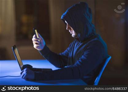 cybercrime, hacking and technology concept - male hacker with laptop computer and smartphone in dark room. hacker with laptop and smartphone in dark room. hacker with laptop and smartphone in dark room