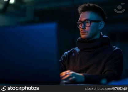 cybercrime, hacking and technology concept - young man in glasses writing code on laptop in dark room. young man in glasses with laptop in dark room