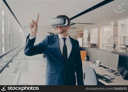 Cyberspace experience at work. Happy man office worker wearing vr goggles, pointing with finger at objects in digital world, businessman in 3d glasses interacting with virtual reality, dressed in suit. Happy man office worker wearing vr goggles, pointing with finger at objects in digital world