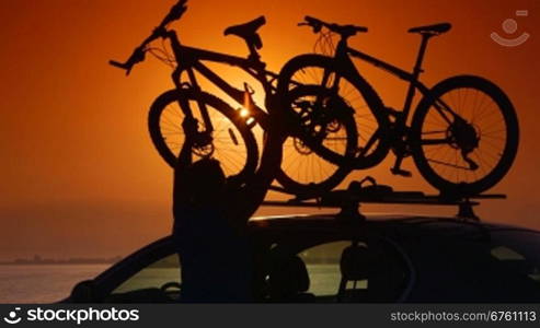 Cyclist attaching bikes to car roof carrier on summer beach at sunset