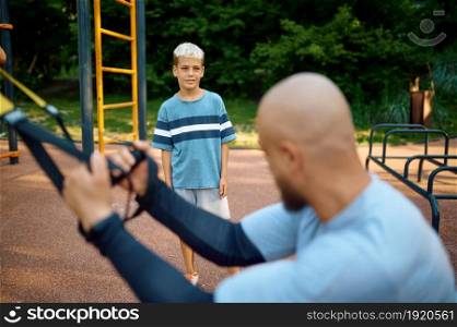 Dad and son doing exercise with ropes, sport training on playground outdoors. The family leads a healthy lifestyle, fitness workout in summer park. Dad and son doing exercise with ropes, playground