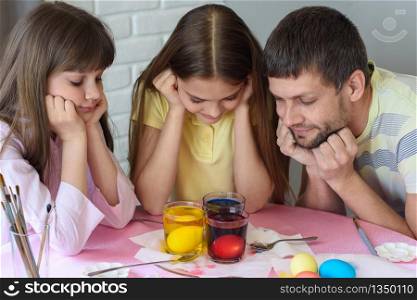 Dad and two daughters are watching how the eggs are painted in glasses with dyes