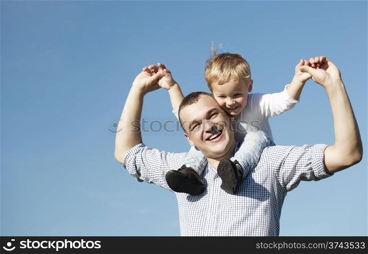 Dad giving his young son a piggy back ride as they both laugh with pleasure and enjoyment, low angle against a clear blue summer sky with copyspace