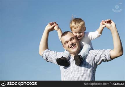 Dad giving his young son a piggy back ride as they both laugh with pleasure and enjoyment, low angle against a clear blue summer sky with copyspace