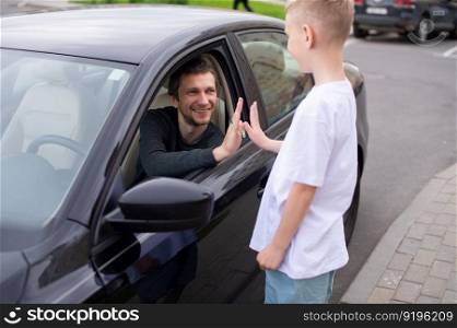 Dad smiles at his son, the child meets the father, a black car, a meeting in the city. to come home, to say goodbye near the car, to see off to work, a boy in a white T-shirt, his father&rsquo;s smile, an open window. A cute child accompanies a happy father to work. Dad is sitting in the car. Give five
