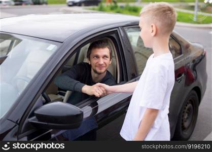 Dad smiles at his son, the child meets the father, a black car, a meeting in the city. to come home, to say goodbye near the car, to see off to work, a boy in a white T-shirt, his father&rsquo;s smile, an open window. A cute child shakes hands with a happy father . Dad is sitting in the car and smiling
