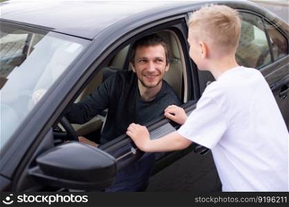 Dad smiles at his son, the child meets the father, a black car, a meeting in the city. to come home, to say goodbye near the car, to see off to work, a boy in a white T-shirt, his father&rsquo;s smile, an open window. A cute child accompanies a happy father to work. Dad is sitting in the car