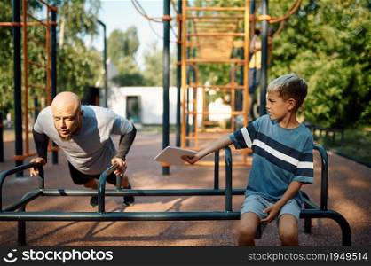 Daddy and son on exercise machine, sport training on playground outdoors. The family leads a healthy lifestyle, fitness workout in summer park. Daddy and son on exercise machine, sport training