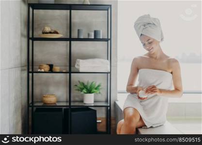 Daily routine, wellness and hygiene concept. Pleased young slim young European woman wrapped in bath towel applies body cream, has healthy skin, poses in cozy bathroom, takes care of herself.