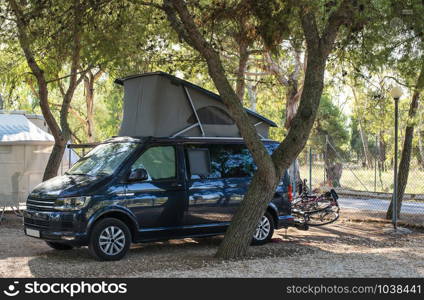 Dark blue camper caravan on camping. Morning sun. Motorhome on campsite and trees around it.