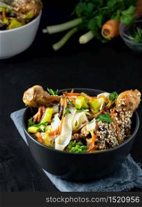 Dark bowl with noodles is located on a dark background. Asian-style lunch, noodles with chicken in teriyaki sauce, vegetables, spices and microgreens, ingredients for noodles on the table. Noodles with vegetables, chicken and teriyaki sauce, closeup.
