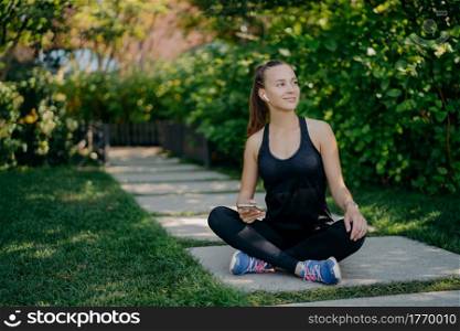 Dark haired young European woman sits crosses legs looks aside with satisfied expression uses smartphone has break after cardio training dressed in active wear and sneaker reads text message