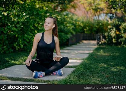 Dark haired young European woman sits crosses legs looks aside with satisfied expression uses smartphone has break after cardio training dressed in active wear and sneaker reads text message