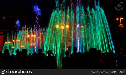 Dark silhouettes of people around the fountainin the late evening, multi-colored neon lights illuminate the jets of a powerful urban singing fountain.. Multi-colored neon lights illuminate the jets of a powerful urban singing fountain in the late evening.