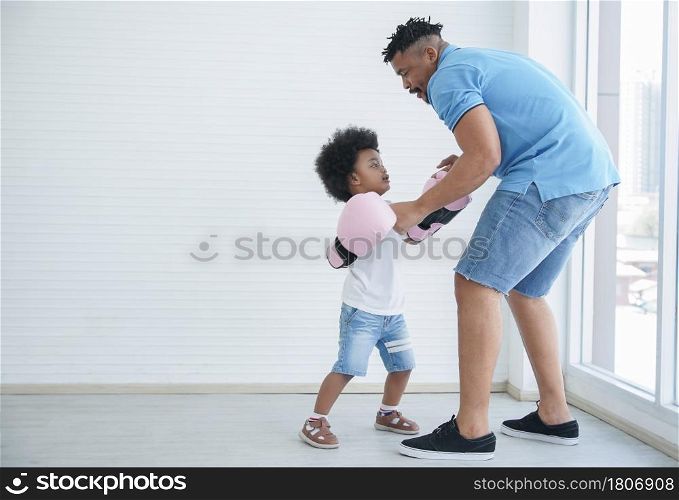 Dark skinned African father and little kid son playing with boxing gloves. Young dad with beard and child boy working out and fun together on white background at home near window