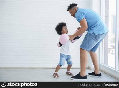 Dark skinned African father and little kid son playing with boxing gloves. Young dad with beard and child boy working out and fun together on white background at home near window