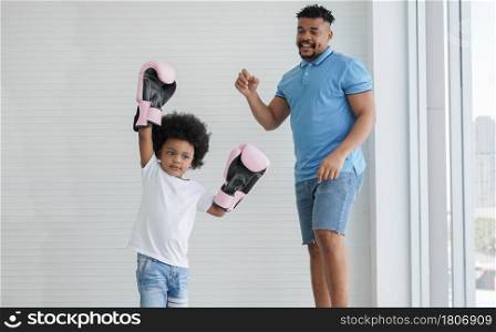 Dark skinned African father and little kid son playing with boxing gloves. Active child boy raising two hands to win his young dad and fun together on white background at home