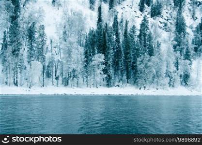 Dark snow forest near winter river. Lake shore is covered with ice and frost. Light frosty haze over water from cold.