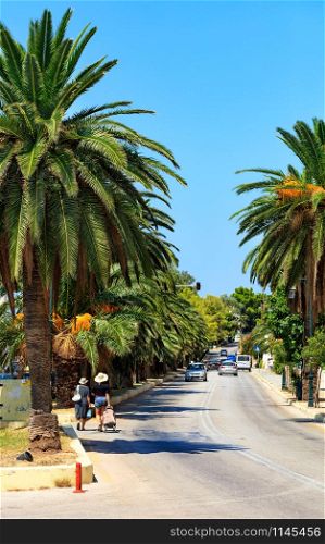 Date palms with fiery orange fruits grow along the roadway, forming a beautiful alley, cars drive along the road, visiting tourists walk and violating the rules of the road in Loutraki, Greece, image with copy space.. Along the road, an alley of date palm trees grows, cars go along the road, females walk, violating traffic rules, a vertical image.