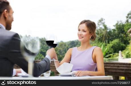 dating, celebration and valentines day concept - smiling young couple clinking glasses of non-alcoholic red wine and looking at each other at restaurant over summer background. young couple with glasses of wine at restaurant