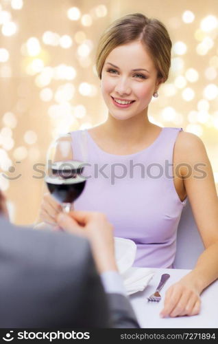 dating, celebration and valentines day concept - smiling young woman clinking glass of non-alcoholic red wine with her man at restaurant over festive lights on beige background. woman drinking wine with her man at restaurant