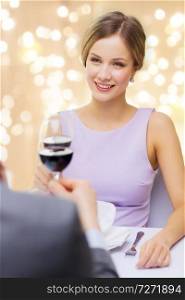dating, celebration and valentines day concept - smiling young woman clinking glass of non-alcoholic red wine with her man at restaurant over festive lights on beige background. woman drinking wine with her man at restaurant