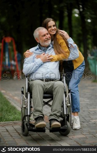 Daughter and disabled father in wheelchair, happy family walking in park. Paralyzed people and disability, handicap overcoming. Handicapped male person and young female guardian in public place. Daughter and disabled father, family in park
