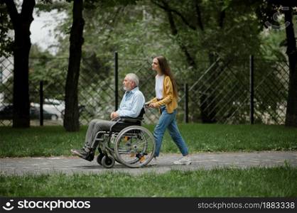 Daughter and disabled father in wheelchair on a walking path, happy family in park. Paralyzed people and disability, handicap overcoming. Handicapped male person and female guardian in public place. Daughter and disabled father on a walking path