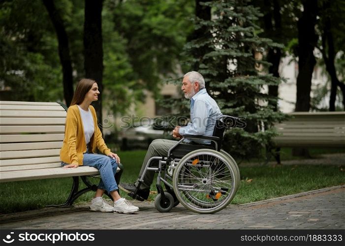 Daughter and disabled father in wheelchair resting on a bench. Paralyzed people and disability, handicap overcoming. Handicapped male person and young female guardian leisure in public place. Daughter and disabled father resting on a bench