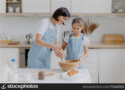 Daughter and mother love cooking, dressed in striped aprons, prepare delicious dinner, use different ingredients, have kitchen routine, stand at home. Children, motherhood, culinary concept.