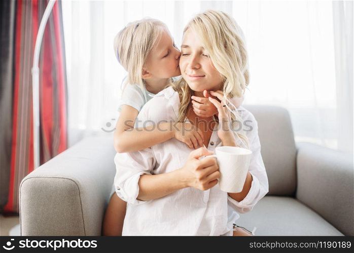 Daughter kisses mom sitting on the couch with a cup of coffee. Parent feeling, togetherness, happy family
