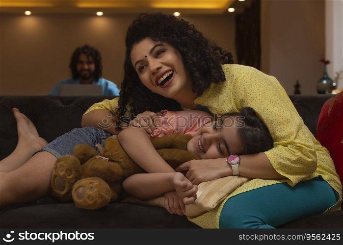 Daughter lying on her mother lap on sofa and father watching them from a distance