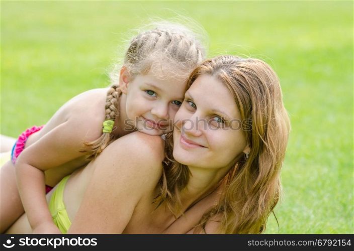 Daughter lying on mother's back happily hugs her, against the background of green grass