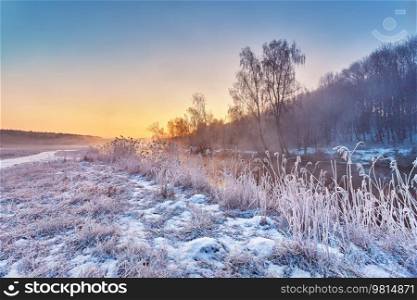 Dawn on winter riverbank. Frost on grass, cane. Snowy rural road. January fog. Forest river sunrise. Cold Weather landscape, reflection in water. Europe rural scene