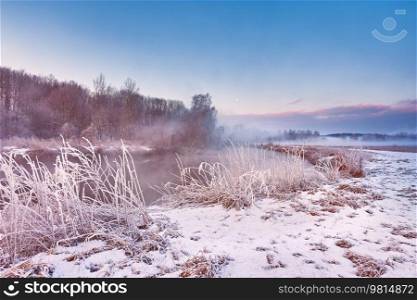 Dawn on winter riverbank. Frost on grass, cane. Snowy rural road. January fog. Forest river sunrise. Cold Weather landscape, reflection in water. Europe rural scene