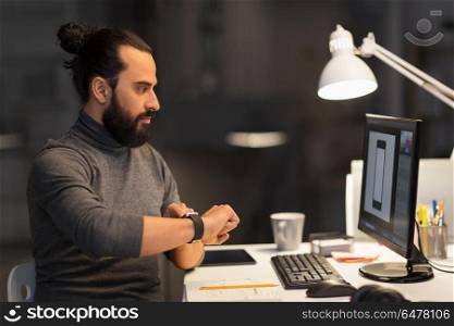 deadline, technology and people concept - creative man or graphic designer with smartwatch and computer working late at night office. designer with smartwatch and computer at office. designer with smartwatch and computer at office