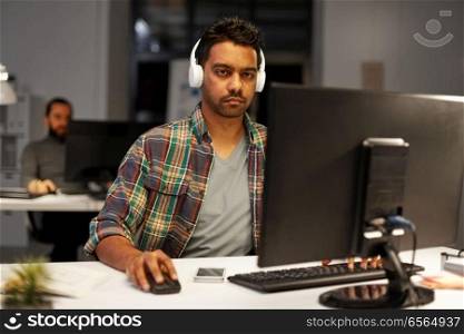 deadline, technology and people concept - creative man with headphones and computer listening to music and working at night office. creative man with headphones working at office