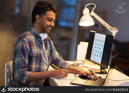 deadline, technology and people concept - creative man with laptop computer working at night office and writing to notebook. creative man with laptop working at night office