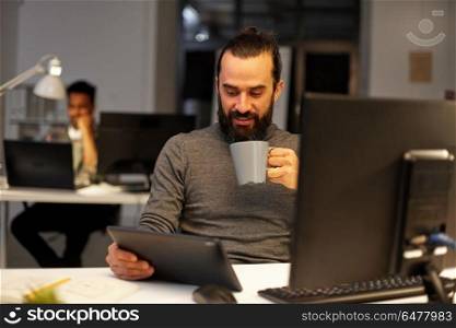 deadline, technology and people concept - creative man with tablet pc computer working late at night office and drinking coffee. man with tablet pc drinking coffee at office. man with tablet pc drinking coffee at office