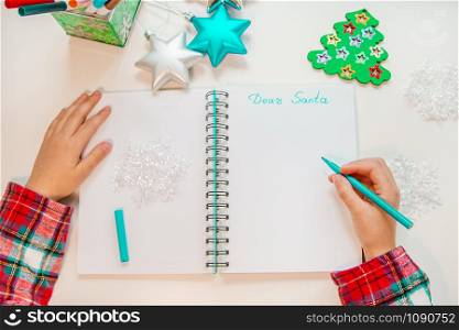 Dear Santa letter, Christmas card. A child holding a pen writes on a white sheet on a wooden background with New Year&rsquo;s decor. Childhood dreams about gifts. New Year concept.. Dear Santa letter, Christmas card. A child holding a pen writes on a white sheet on a wooden background with New Year&rsquo;s decor.