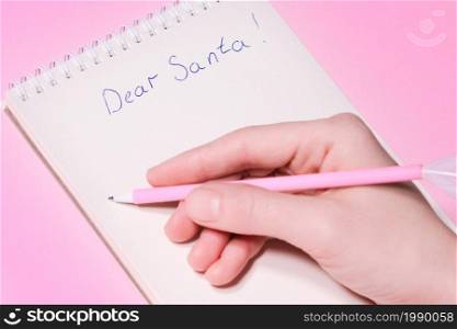 Dear Santa letter concept. Hand holding pink pen decorated with feather and writing in Notepad letter to Santa Claus on pink background. Dear Santa letter concept. Hand holding pink pen decorated with feather and writing in Notepad letter to Santa Claus on pink background.
