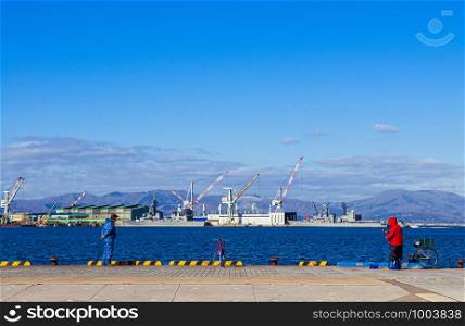 DEC 2, 2018 Hakodate, JAPAN -Hakodate blue harbour bay with people fishing and industrial port with large cranes and ship mountain view background, Distant view.