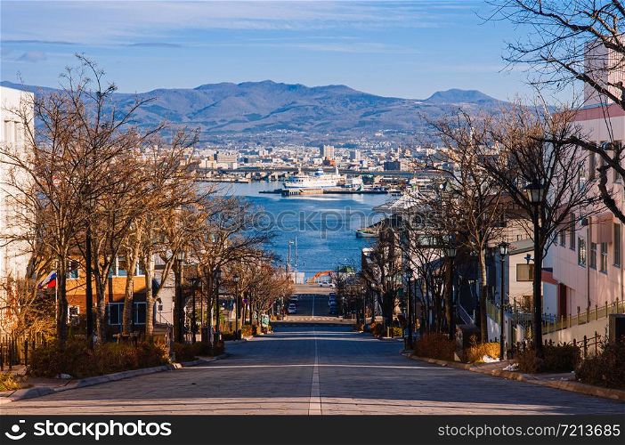 DEC 2, 2018 Hakodate, JAPAN -Motomachi slope Street with leafless tree along both sides with people crossing road Hakodate bay in background with blue winter sky.