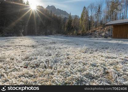 December scenery with grass covered by snow and ice, a bright sun, forest and a wooden cabin, near Garmisch-Partenkirchen, Germany.