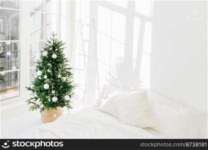 Decorated festive Christmas tree in white classic bedroom interior with soft bed. Winter home interior. Cozy season. New Year, celebration