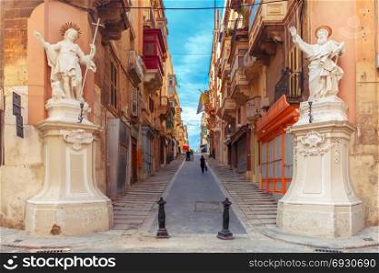 Decorated street in old town of Valletta, Malta. The traditional Maltese street stairs with corners of houses, decorated with statues of saints St. John and St. Paul and building with colorful balconies in Valletta, Capital city of Malta